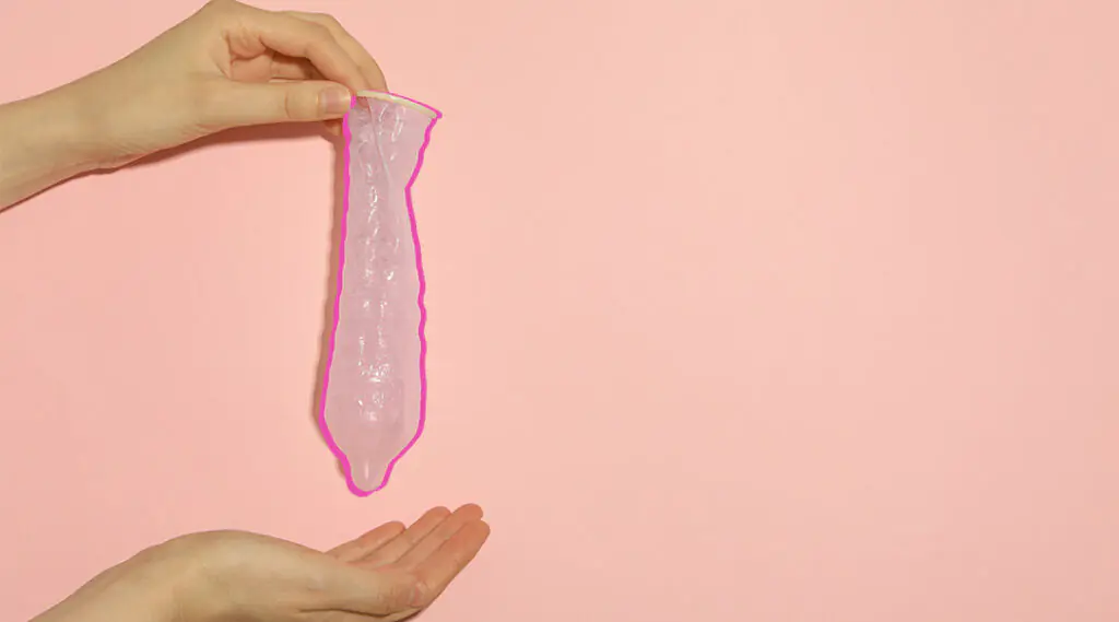 Preservativo sostenido entre dos manos sobre fondo rosa.