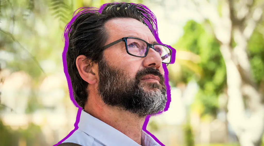Hombre adulto con barba y gafas observa hacia un lado en un entorno exterior.