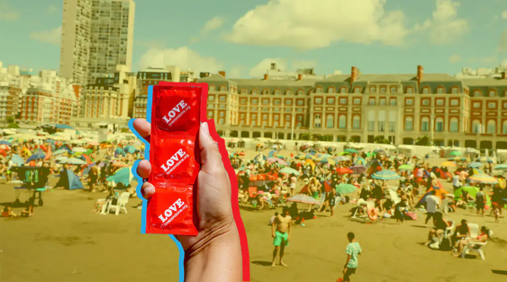 Mano sostiene preservativo rojo de la marca LOVE frente a una playa concurrida.