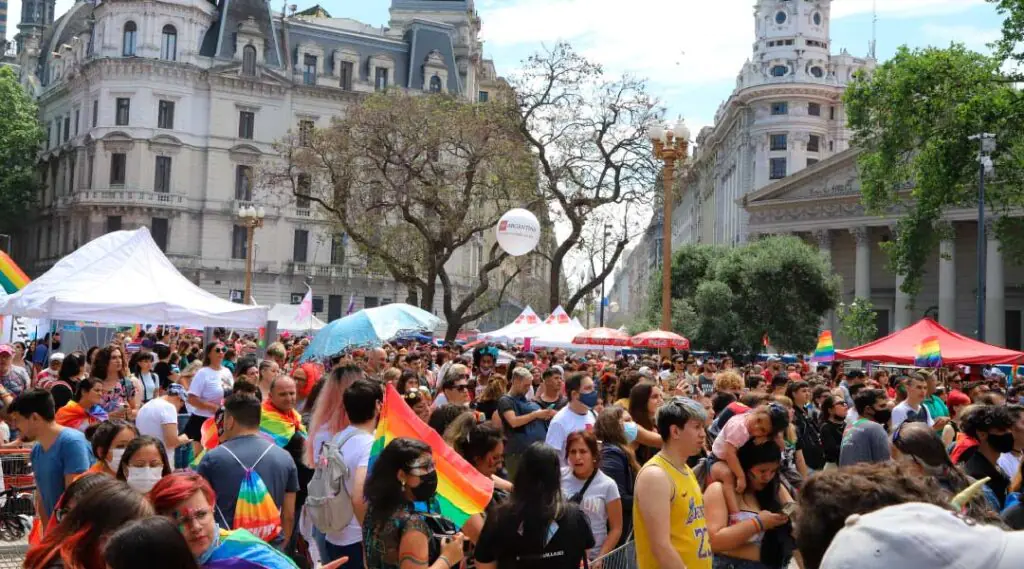Personas reunidas en evento del orgullo LGBTQ+ con banderas arcoíris y stands en espacio público.