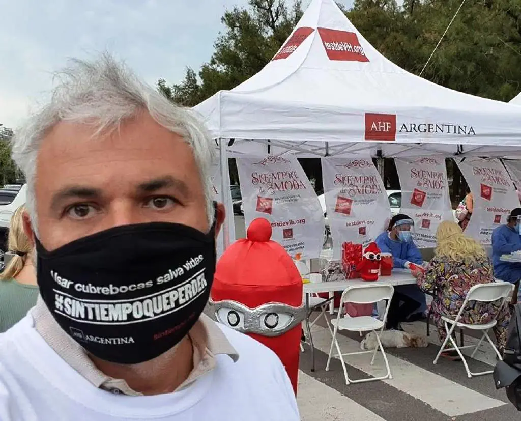 Persona con barbijo toma una selfie frente a un stand de AHF Argentina con carpa y mesas de atención.